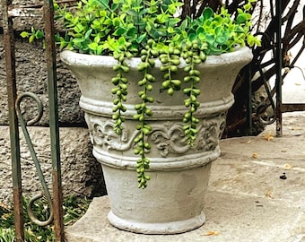 May include: A light gray concrete planter with a flared rim and a decorative embossed scroll pattern around the center. The planter is filled with vibrant green artificial eucalyptus foliage and trailing string of pearls succulent stems.