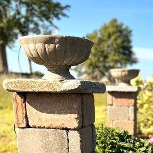 May include: A weathered stone urn with a fluted exterior sits atop a square stone pedestal constructed from stacked rectangular blocks. The urn features a wide, shallow bowl shape with a pedestal base, showcasing a textured, aged gray finish.