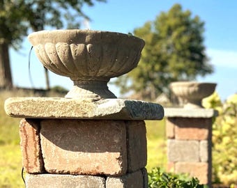 May include: A weathered stone urn with a fluted exterior sits atop a square stone pedestal constructed from stacked rectangular blocks. The urn features a wide, shallow bowl shape with a pedestal base, showcasing a textured, aged gray finish.