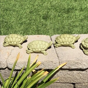 May include: Four small, light green turtle figurines are arranged on a stone wall. The turtles have detailed shell patterns and are positioned in a row. The background features green grass and some plants.