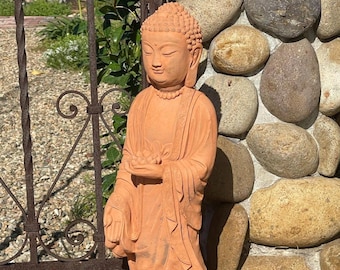 May include: A terracotta-colored standing Buddha statue featuring a serene facial expression, curled hair, and draped robes. The figure holds a small lotus flower in one hand while the other hand is extended downward in a gesture of offering.