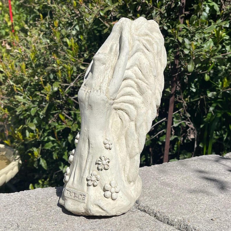 May include: White concrete garden statue of a woman with her head tilted back and her arms raised. The statue has a floral design and the word "TRIBUTE" is inscribed on the base.