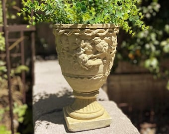 May include: A beige stone-colored urn planter featuring a relief design of birds and foliage on the exterior. The vessel has a flared rim with a braided texture, a tapered body, and a square base, filled with a vibrant green leafy plant.