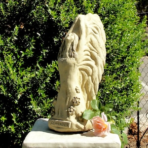 May include: A beige stone sculpture of a figure with large wings and long hair, standing with arms raised. The base features the word REMEMBER in raised letters, and the figure is decorated with small flower carvings on the lower portion.
