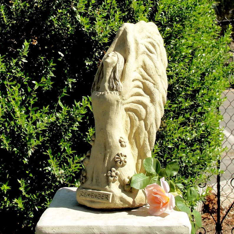 May include: A beige stone sculpture of a figure with large wings and long hair, standing with arms raised. The base features the word REMEMBER in raised letters, and the figure is decorated with small flower carvings on the lower portion.