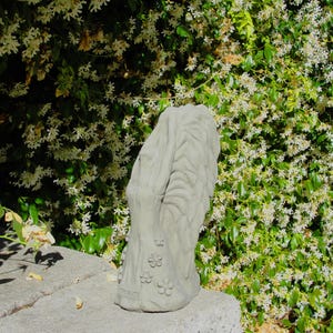 May include: A light gray concrete garden sculpture featuring a vertical, textured tree trunk design with carved floral accents at the base. The object has a weathered, matte finish and stands upright with a rounded top and a wide, stable base.