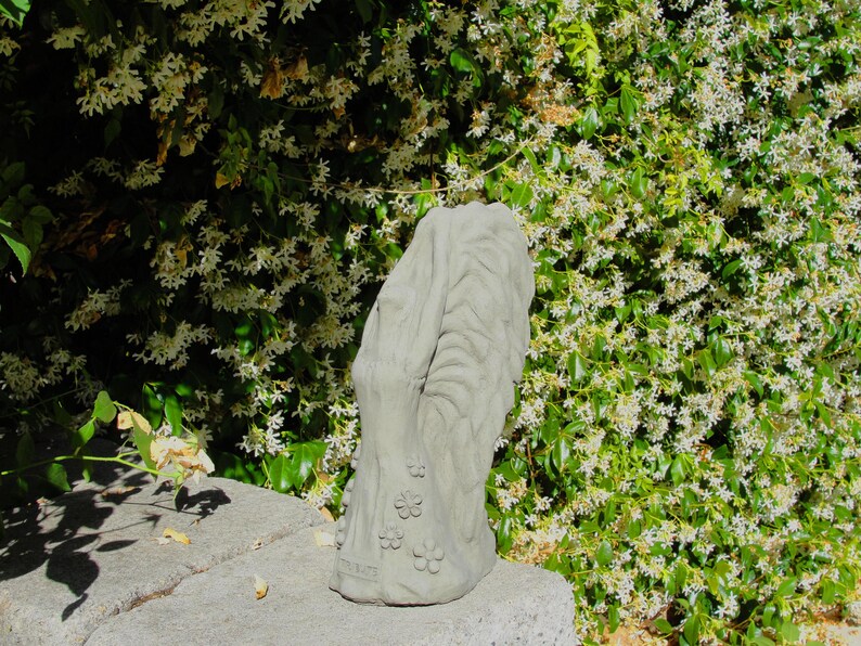 May include: A light gray concrete garden sculpture featuring a vertical, textured tree trunk design with carved floral accents at the base. The object has a weathered, matte finish and stands upright with a rounded top and a wide, stable base.