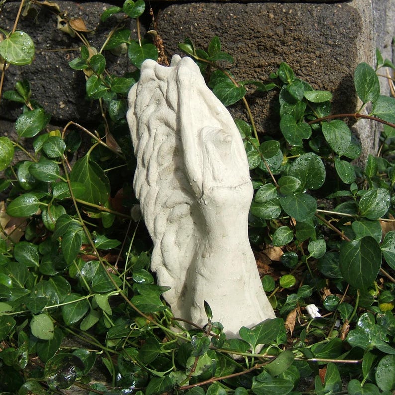 May include: A light-colored, off-white stone sculpture featuring a textured, vertical form with a braided or woven appearance on one side and a smooth, elongated surface on the other, standing upright among green ivy leaves and dark stone.