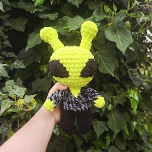 Puede incluir: Muñeco alienígena de ganchillo hecho a mano en verde neón brillante, negro y gris. El alienígena tiene ojos negros grandes, una camisa estampada gris y negra y pantalones negros. El muñeco mide unos 20 cm de alto.