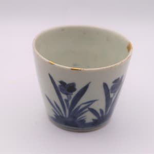 May include: A white ceramic cup with a blue floral design. The cup has a slightly flared shape and a gold rim. The blue floral pattern includes stylized flowers and leaves. The cup is likely vintage or antique.
