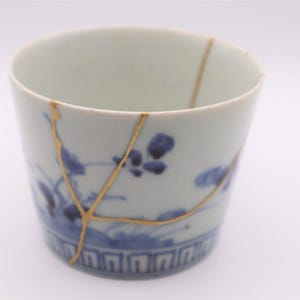 Puede incluir: Una taza de cerámica blanca y azul con un diseño floral y reparación kintsugi dorada. La taza tiene forma cilíndrica y un borde decorativo en la base. La reparación kintsugi resalta las grietas.