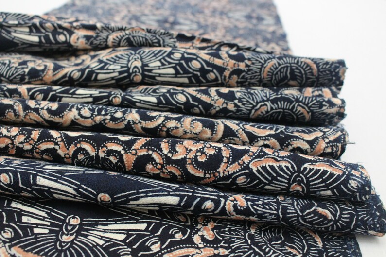 May include: A stack of folded fabric with a repeating pattern of white and tan designs on a navy blue background. The fabric appears to be cotton or a similar material, with visible folds and edges.