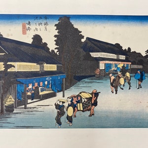 May include: A Japanese woodblock print depicting a street scene with people walking and carrying goods. The print is in shades of blue, green, and brown, with a light blue sky in the background. The text in the image reads "天保三年", which translates to "Third year of Tenpo."