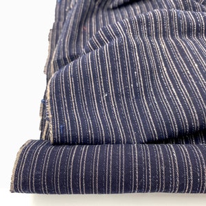 May include: Close-up of folded navy blue fabric with vertical white stripes. The fabric is textured and appears to be a woven material. The edges of the fabric are visible, showing the raw, unfinished edges. The fabric is layered, creating depth and dimension.