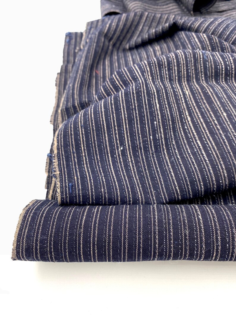 May include: Close-up of folded navy blue fabric with vertical white stripes. The fabric is textured and appears to be a woven material. The edges of the fabric are visible, showing the raw, unfinished edges. The fabric is layered, creating depth and dimension.