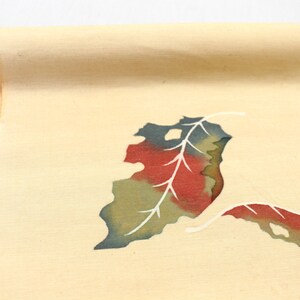 May include: Rolled-up beige fabric with a leaf design. The leaves are colored with red, green, and white accents. The fabric is likely used for decorative purposes or crafting.