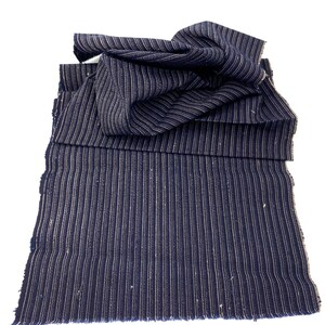 May include: Navy blue fabric with vertical white stripes. The fabric is folded and layered, with a frayed edge visible at the bottom. The material appears to be a woven textile, possibly for clothing or home decor.