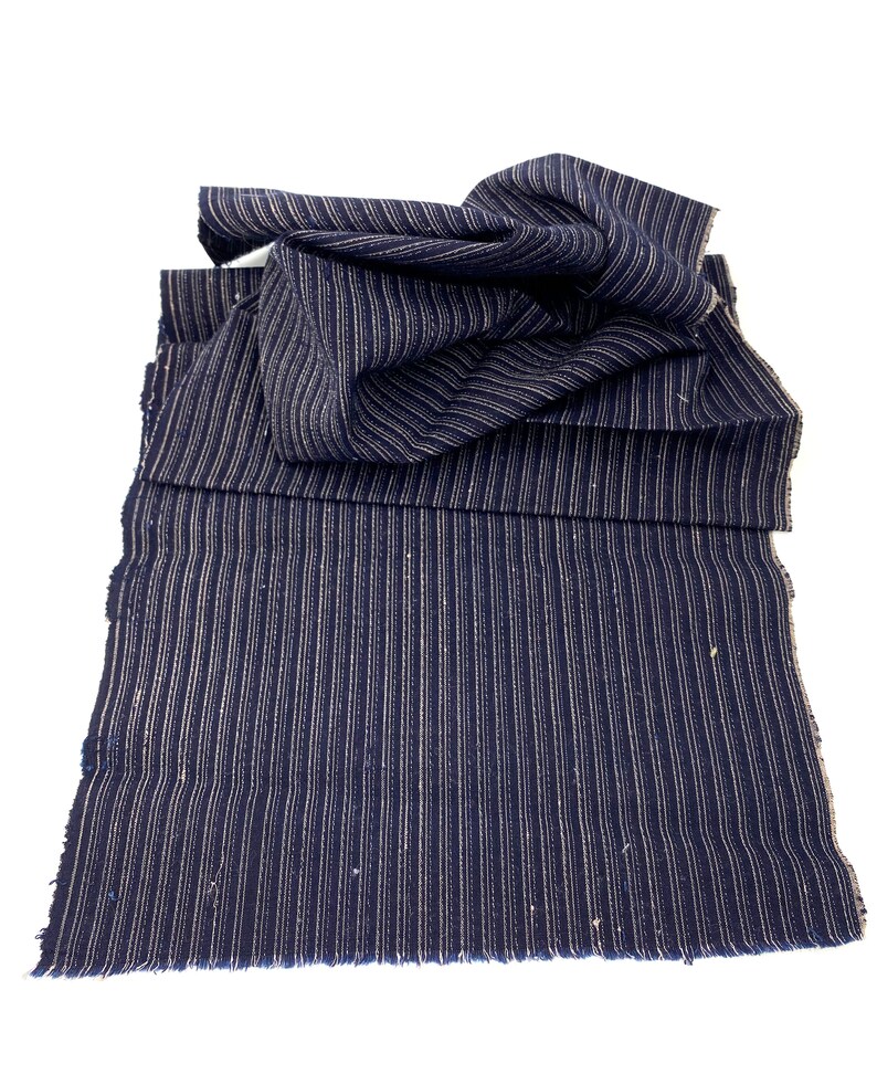 May include: Navy blue fabric with vertical white stripes. The fabric is folded and layered, with a frayed edge visible at the bottom. The material appears to be a woven textile, possibly for clothing or home decor.