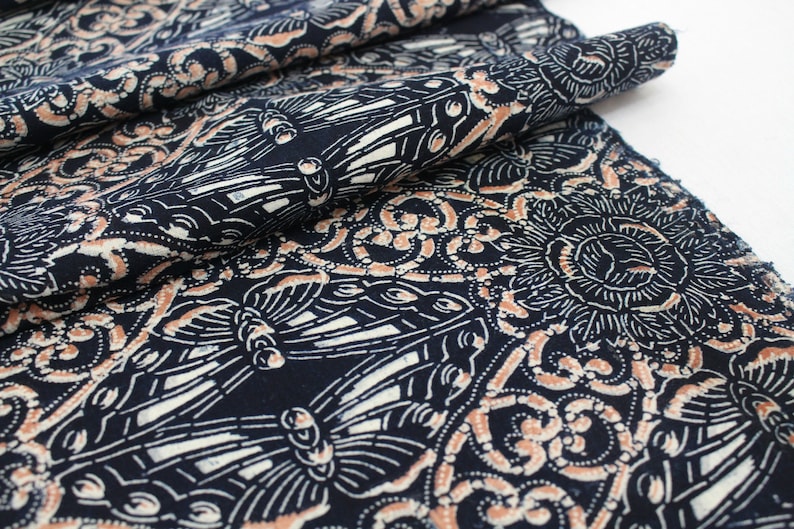 May include: Dark blue fabric with a repeating pattern of butterflies, floral designs, and swirls in white and peach. The fabric is folded, showing the detailed print and texture. Ideal for sewing projects.