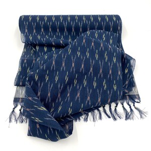 May include: A navy blue scarf with a repeating pattern of vertical lines in yellow, tan, and white. The scarf has a fringed edge and is made of a lightweight fabric. The scarf is folded and draped on a white surface.