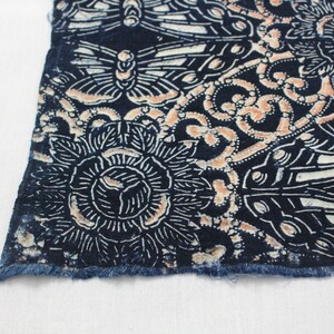 May include: Close-up of a section of fabric with a detailed pattern. The design features stylized flowers and butterflies in white and peach against a dark blue background. The edge of the fabric has a frayed, unfinished look.