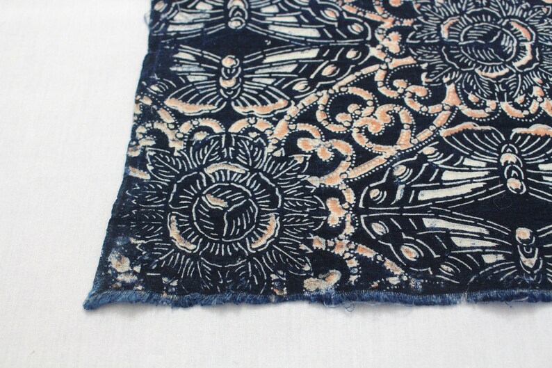 May include: Close-up of a section of fabric with a detailed pattern. The design features stylized flowers and butterflies in white and peach against a dark blue background. The edge of the fabric has a frayed, unfinished look.