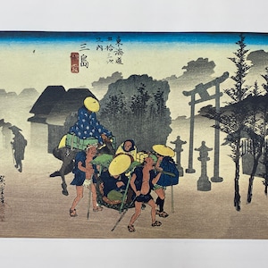 May include: A Japanese woodblock print depicting a group of people traveling through a misty forest. The print is in shades of blue, gray, and brown, with a touch of yellow. The people are carrying various items, including a large basket and a small animal. The print is titled "東海道 三十三  之内  三島", which translates to "The Tokaido Road, 33 Stations, Mishima."