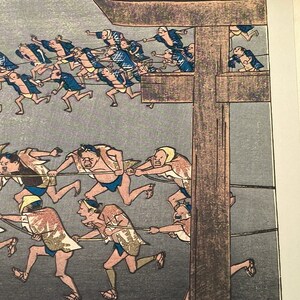 May include: A woodblock print showing figures pulling ropes, set against a muted grey backdrop. The people are dressed in traditional Japanese attire, with some wearing blue jackets and others patterned garments. The print has a historical feel.