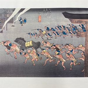 May include: Japanese woodblock print showing a nighttime scene with figures pulling a horse. The print features a building on the left, a large gate on the right, and numerous figures running with ropes. The colour palette includes shades of blue, grey, and brown.