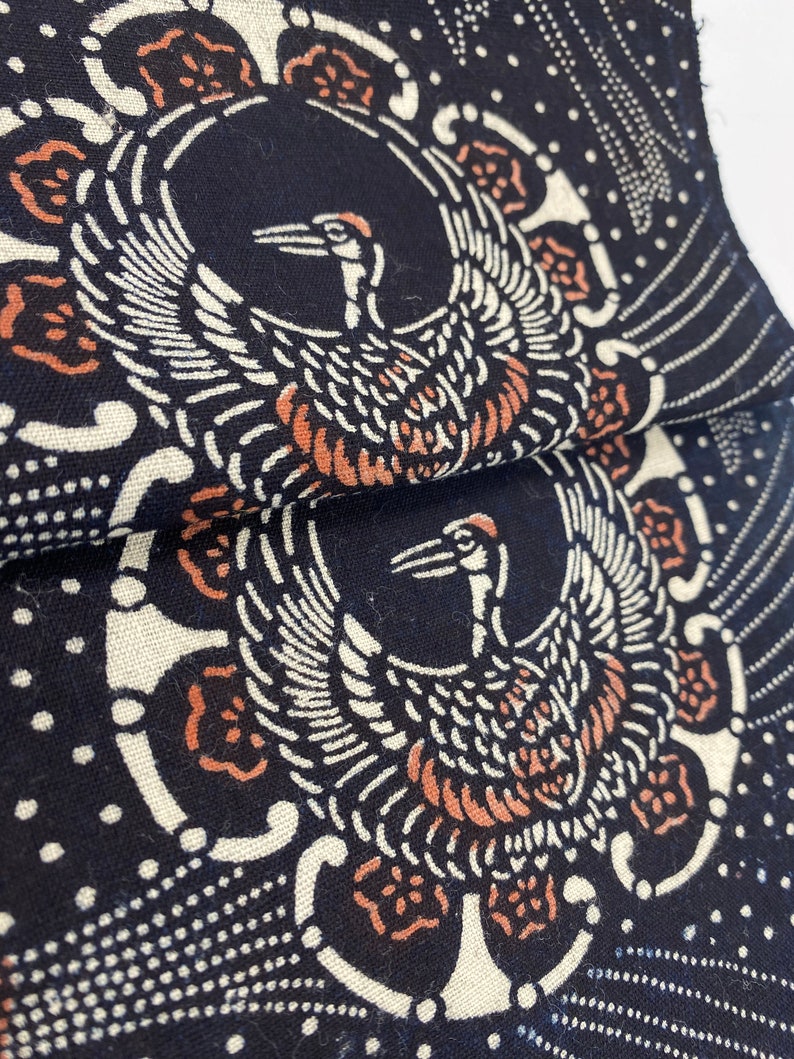 Puede incluir: Tela negra con un dise&ntilde;o blanco y naranja de dos grullas con las alas extendidas. La tela tiene un patr&oacute;n repetitivo de peque&ntilde;os puntos blancos.