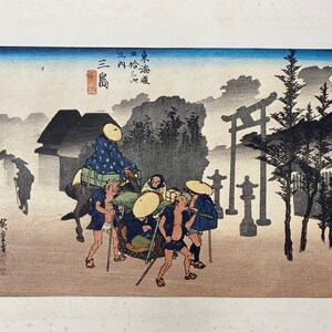 May include: A Japanese woodblock print depicting a group of people traveling through a misty forest. The people are carrying a palanquin, and there is a torii gate in the background. The print is in shades of gray, blue, and brown.