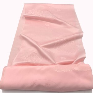 May include: A pale pink, silky fabric with a floral pattern. The fabric is folded and draped, revealing a subtle sheen. The design includes flowers and other decorative elements. The fabric is likely used for clothing or home decor.