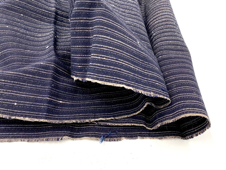 May include: Close-up of a folded fabric with a navy blue background and white horizontal stripes. The fabric has a raw, unfinished edge. The material is bunched up, showing the texture and pattern of the textile.