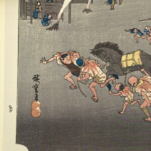 May include: Japanese woodblock print showing a dynamic scene with running figures and a horse-drawn carriage. The print uses a subdued colour palette of grey, brown, and blue. The artwork displays traditional Japanese clothing and architectural details.