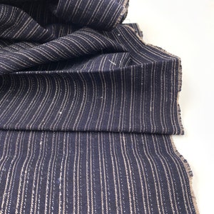 May include: Dark navy blue fabric with vertical, thin, light beige stripes. The fabric is folded and draped, showing the texture and pattern. The edges are unfinished, revealing the raw weave of the material. Ideal for sewing projects.
