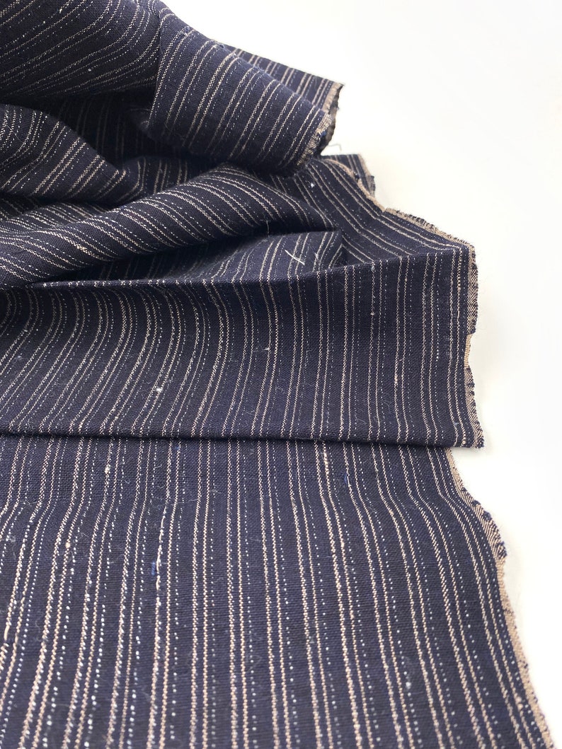 May include: Dark navy blue fabric with vertical, thin, light beige stripes. The fabric is folded and draped, showing the texture and pattern. The edges are unfinished, revealing the raw weave of the material. Ideal for sewing projects.