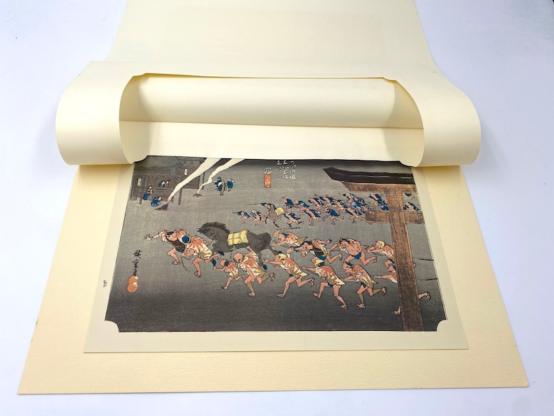 May include: A Japanese woodblock print showing a group of people running, pulling a horse-drawn cart. The print features a dark, stormy sky and traditional Japanese architecture. The print is framed by a cream-coloured mount.