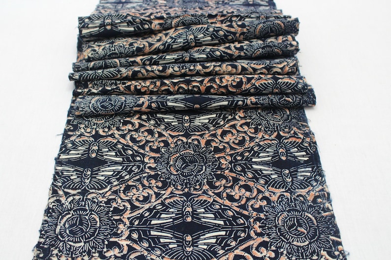May include: Folded fabric with a repeating pattern of stylized butterflies and floral elements in navy blue, cream, and peach. The fabric is layered, showcasing the detailed design.