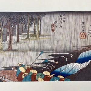May include: A woodblock print depicting a group of people in a boat on a river. The scene is set in a forest with trees on both sides of the river. The people are wearing traditional Japanese clothing and are holding oars. The print is in shades of blue, green, and brown.