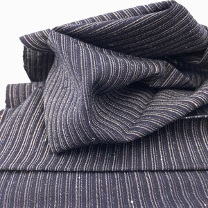 May include: Close-up of navy blue fabric with vertical white stripes. The fabric is folded and draped, showcasing its texture and pattern. The stripes are evenly spaced, creating a classic and versatile design. The fabric appears to be a medium weight.