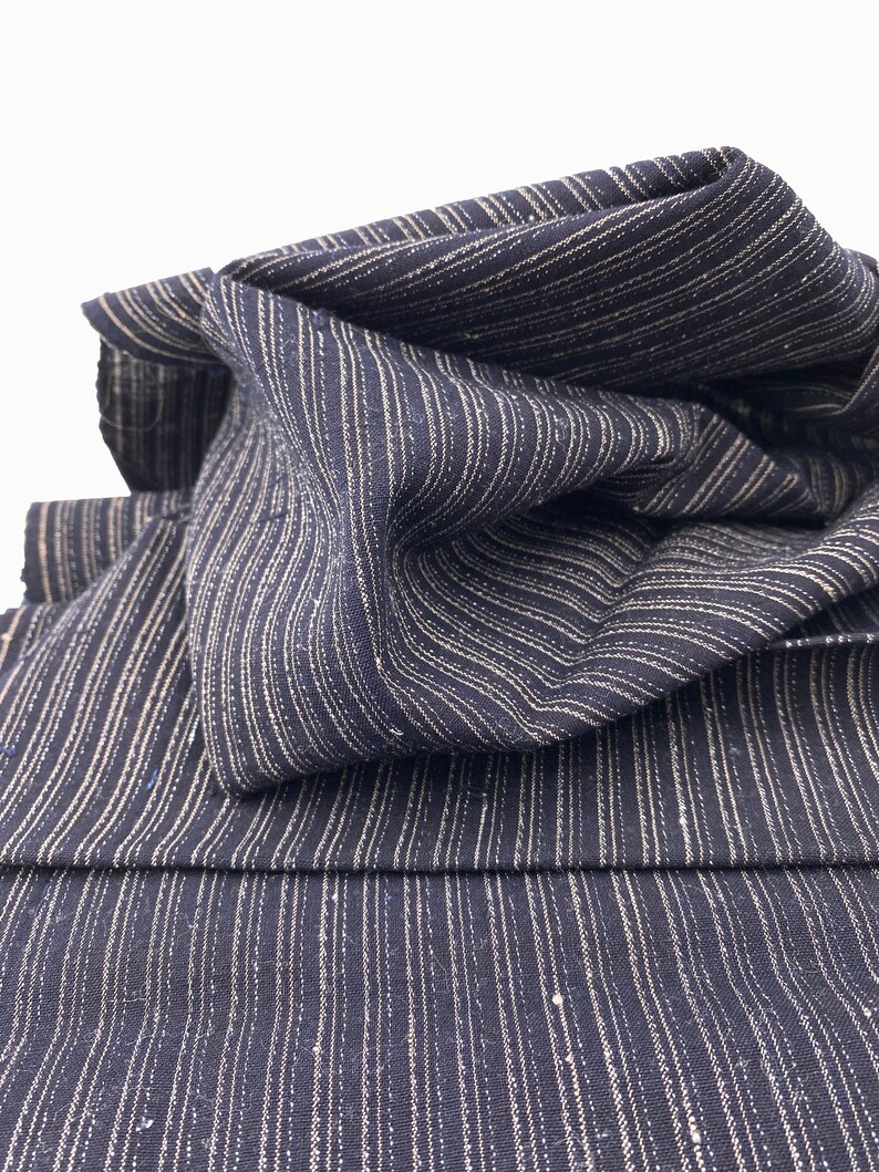 May include: Close-up of navy blue fabric with vertical white stripes. The fabric is folded and draped, showcasing its texture and pattern. The stripes are evenly spaced, creating a classic and versatile design. The fabric appears to be a medium weight.