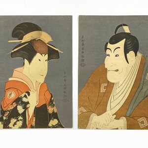 May include: Two woodblock prints of Japanese actors in traditional clothing. The print on the left features a woman with a black wig and a red and orange kimono. The print on the right features a man with a black wig and a brown kimono.