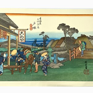 May include: A Japanese woodblock print depicting a scene of travelers on a road. The print features a group of people on horseback, a man walking across a bridge, and a small village in the background. The print is in shades of blue, green, and brown.