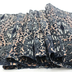 May include: Close-up of a folded fabric with a repeating pattern. The fabric is navy blue with a detailed design in white and peach. The pattern features floral and geometric shapes, creating a textured appearance.