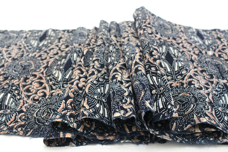 May include: Close-up of a folded fabric with a repeating pattern. The fabric is navy blue with a detailed design in white and peach. The pattern features floral and geometric shapes, creating a textured appearance.