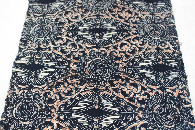 May include: A section of fabric with a repeating pattern of stylized flowers, butterflies, and swirling designs in cream, peach, and white against a navy blue background. The fabric has a slightly frayed edge.