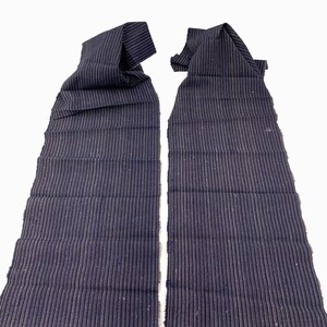 May include: Two pieces of navy blue fabric with vertical, thin, light-colored stripes. The fabric is folded at the top, and the edges are frayed. The background is plain white.