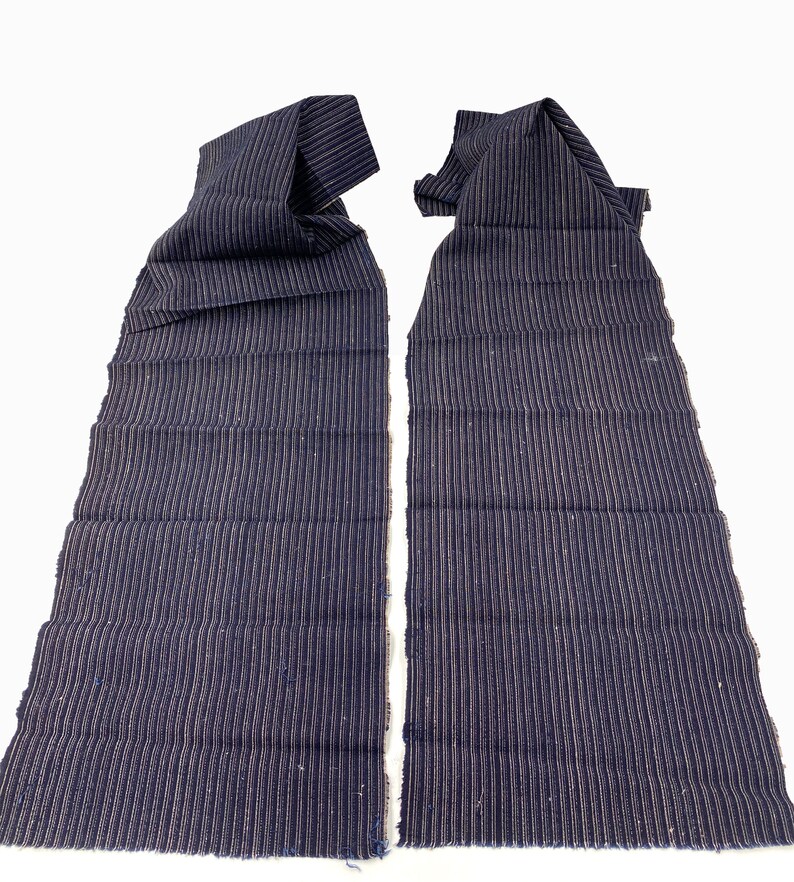 May include: Two pieces of navy blue fabric with vertical, thin, light-colored stripes. The fabric is folded at the top, and the edges are frayed. The background is plain white.