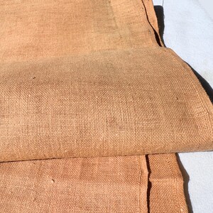 May include: Close-up of folded, textured fabric in a warm, earthy tone. The fabric appears to be linen or a similar natural fiber, with visible weave detail. A section of white fabric is visible in the background.