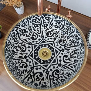 Puede incluir: Un lavabo redondo de cerámica con un patrón ornamental en blanco y negro. El lavabo tiene un borde dorado y un desagüe dorado. Los grifos de color cobre son visibles encima del lavabo. Un accesorio decorativo para el baño.